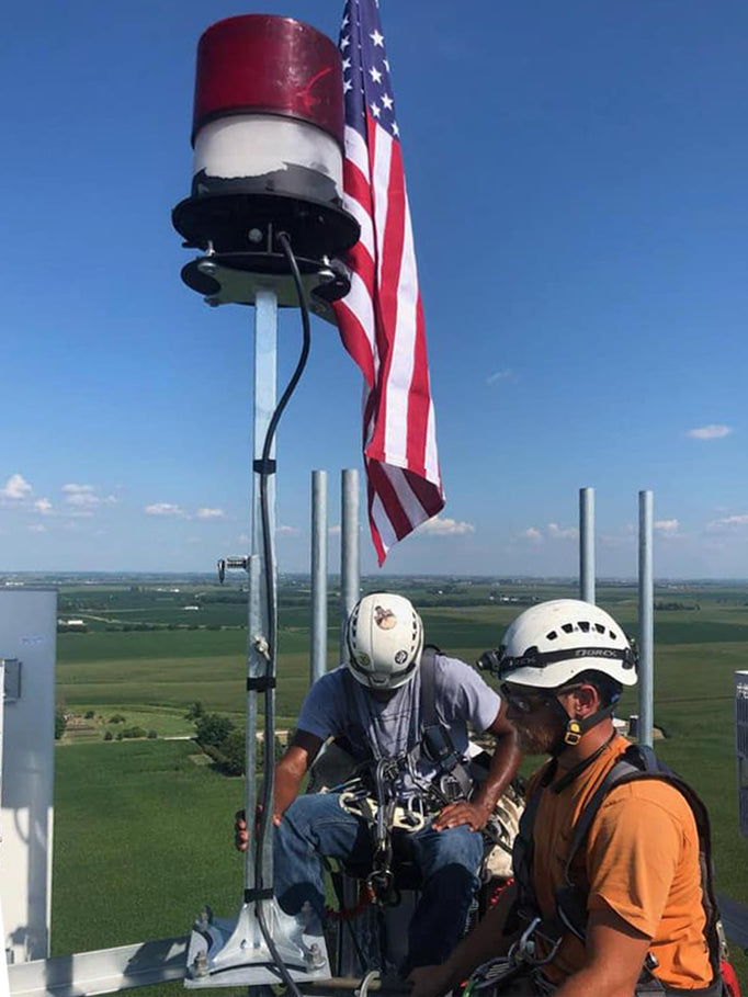 Two Tower Technicians on top of a tower, work on a beacon that is attached to a Remus Tower Beacon Extension, with an American flag beside it.